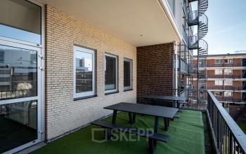 Balkon van een kantoorpand op Kuipersdijk 2-4 in Enschede met groene vloerbedekking en zwarte picknicktafels. Aan de buitenkant zijn bakstenen muren zichtbaar en een wenteltrap naar boven.