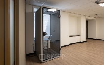 Kantoorruimte op Kuipersdijk 2-4 in Enschede met een moderne werkplekvoorziening en houten vloer. Het interieur is vrij minimalistisch met een stijlvolle telefoniecabine voor privégesprekken.