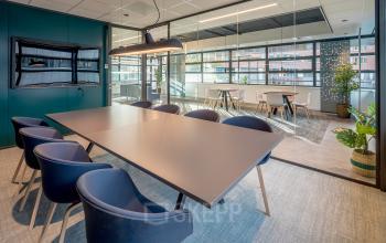 Moderne, lichte kantoorruimte met glazen wanden en vergadertafel, gevestigd op Tielweg 30, Gouda. Ideaal voor zakelijke meetings en interacties, met blauwe stoelen en groene planten voor een uitnodigende sfeer.