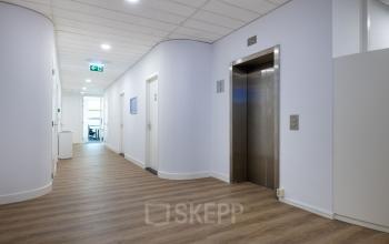 Een lichte gang in een kantoorpand met een houten vloer, witte muren en een moderne lift. De ruimte is goed verlicht en leidt naar verschillende kantoorruimtes.