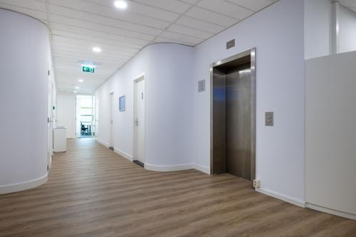 Een lichte gang in een kantoorpand met een houten vloer, witte muren en een moderne lift. De ruimte is goed verlicht en leidt naar verschillende kantoorruimtes.