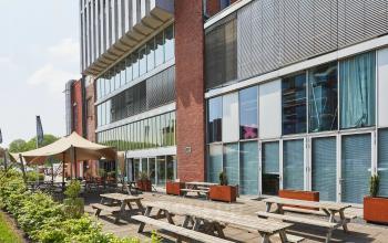 Buitenzijde van een kantoorpand gelegen aan Helperpark 270-298 in Groningen, voorzien van houten picknickbanken en een overkapping op een ruim terras.