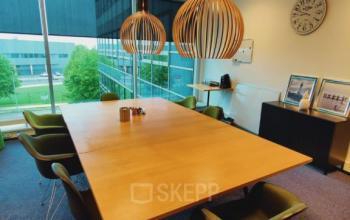 Kantoorruimte huren in kantoorpand Snellius 1 in Heerlen. Vergaderruimte met grote houten tafel, groene stoelen en moderne lampen, met uitzicht op groene omgeving.
