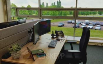 Kantoorpand te huur op Snellius 1 in Heerlen met werkplek, bureaustoel, computerschermen, telefoon, plant en groot raam met uitzicht op parkeerterrein.