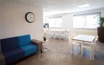 Kantoorpand Steenovenweg 5 in Helmond met modern gemeubileerde ruimte, includes een blauwe en grijze bank, tafels met witte stoelen, en grote ramen voor veel licht.