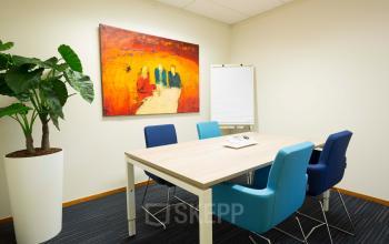 Vergaderruimte met zes stoelen, een houten tafel en grote plant, gelegen op Steenovenweg 5, Helmond. Kleurrijk schilderij aan de muur en flip-over aanwezig. Ideaal voor kantoorruimte huren.