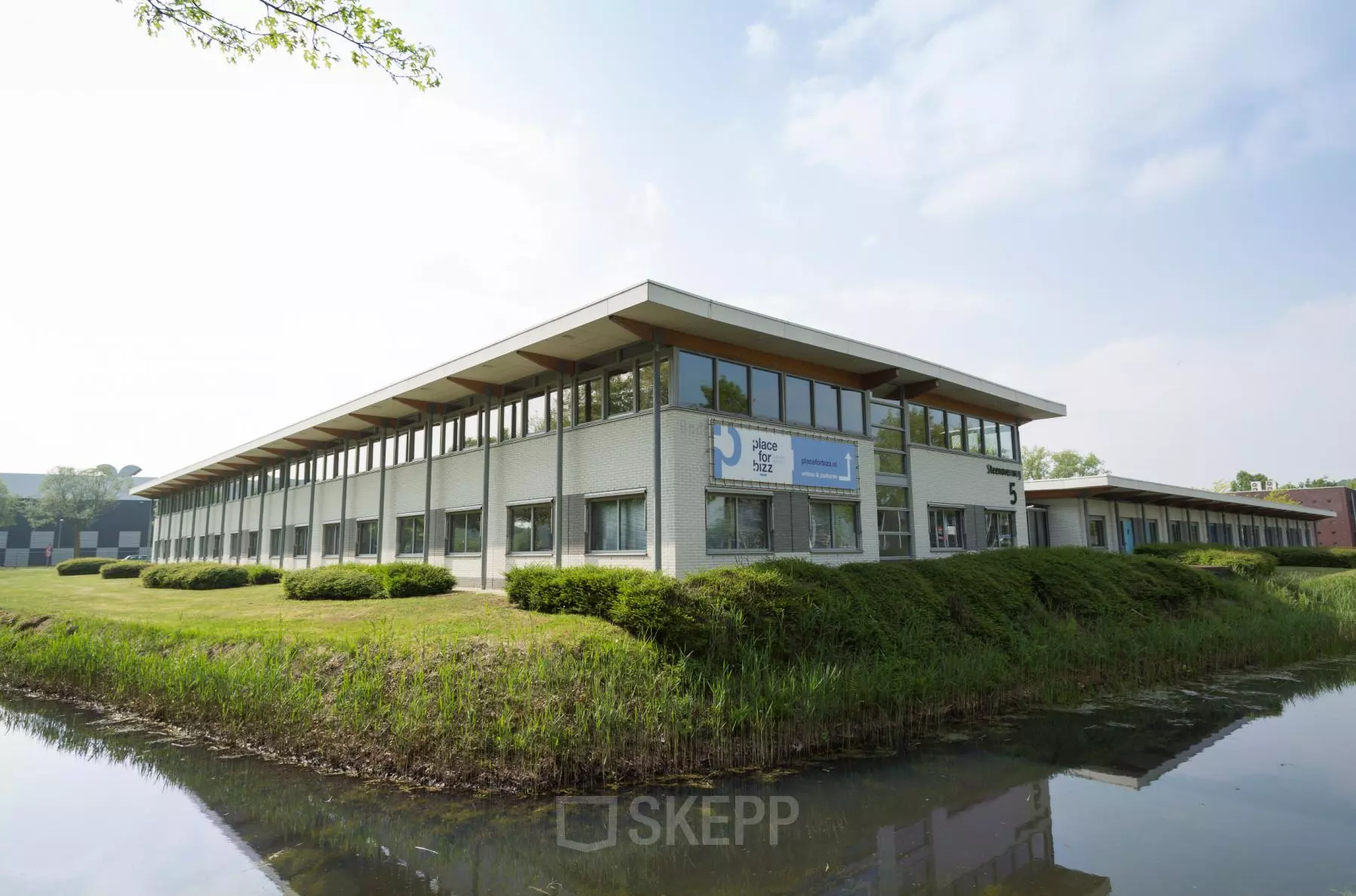 Vooraanzicht van het kantoorpand aan Steenovenweg 5 in Helmond, omringd door groen met waterpartij op de voorgrond.