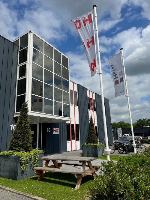 Exterieur van het kantoorpand aan Hamerstraat 10 in Hengelo met moderne gevel en vlaggen, ideaal voor kantoorruimte huren. Voor de ingang staan een picknicktafel en groene beplanting.