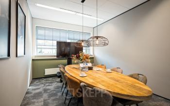 Vergaderruimte in het kantoorpand op Demmersweg 41-47, Hengelo met een houten tafel en stoelen, bloemen, een tv aan de muur en natuurlijke verlichting.