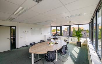 Lichte kantoorruimte op Welbergweg 60, Hengelo met grote ramen, een ovale tafel, stoelen en een whiteboard. Ideaal voor kantoorruimte huren in een kantoorpand. Ruimte is gemeubileerd en klaar voor gebruik.