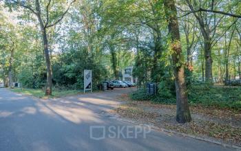 Buitenzicht op het kantoorpand gelegen aan de groene Mozartlaan 25 in Hilversum, omgeven door bomen en met een parkeerplaats vooraan.