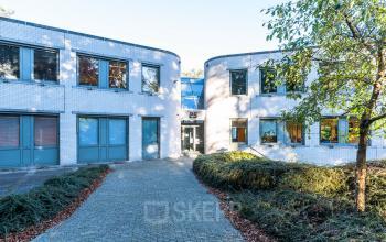 Uitzicht op de buitenkant van het kantoorpand op Mozartlaan 25 in Hilversum, geschikt voor kantoorruimte huren. Het entreepad leidt naar een moderne glazen ingang die omringd is door witte bakstenen muren.