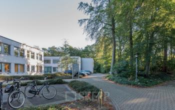 Kantoorpand aan de Mozartlaan 25 in Hilversum, omgeven door bosrijke omgeving. Parkeerplaats met fietsen en auto's zichtbaar.