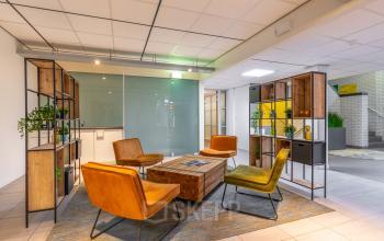 Moderne kantoorlounge in Hilversum met houten rekken, kleurrijke stoelen en een glazen wand, ideaal voor ontspanning tijdens de werkdag. Kantoorruimte huren met verfijnde inrichting.