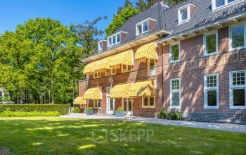Klassiek kantoorpand aan de Utrechtseweg 47 in Hilversum, omgeven door groene beplanting en uitgerust met gele zonwering. Ideaal voor wie kantoorruimte huren wil in een inspirerende omgeving.
