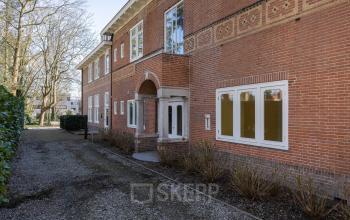 Buitenzijde van kantoorpand aan Utrechtseweg 47, Hilversum. Bakstenen gevel met meerdere grote vensters en sierlijke details. Verscholen in een groene omgeving, ideaal voor kantoorruimte huren.