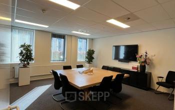 Modern ingerichte vergaderruimte in een kantoorpand aan de Catharina van Renneslaan 8-10, Hilversum, ideaal om te huren. Ruime tafel omringd door comfortabele stoelen, met ramen die natuurlijk licht binnenlaten.