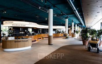 Lichte, moderne kantoorruimte aan Mercuriusplein 1 in Hoofddorp, met grote ramen en houten elementen. Planten zijn strategisch geplaatst voor een groene uitstraling. Mensen lunchen rustig in de gezamenlijke kantine.