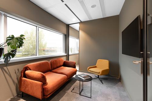 Moderne kantoorruimte met een oranje bank en een gele stoel bij een lage tafel, grote ramen bieden veel natuurlijk licht.