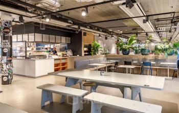 Moderne kantoorruimte in Hoofddorp met gemeubileerde werkplekken, een open keuken en groene accenten. Mensen zien bezig met het bereiden van eten. Ideaal om een kantoorruimte te huren.