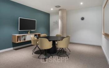 Moderne vergaderruimte met zes beige stoelen rondom een ovale tafel, een muur met een klok en een scherm, ideaal voor kantoorruimte huren op Siriusdreef 17 in Hoofddorp.