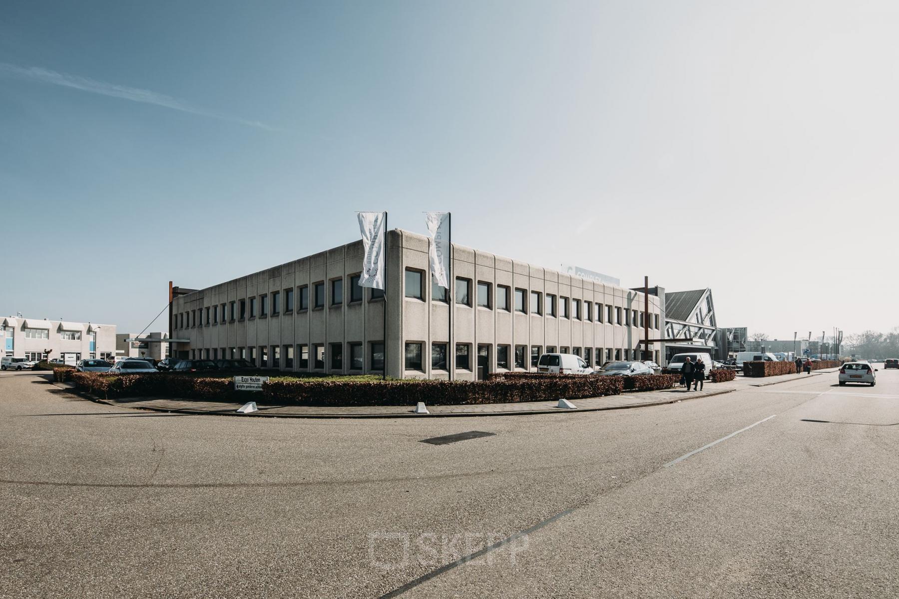 Grijs kantoorpand aan de Meidoornkade 22 in Houten met parkeerplaatsen en vlaggenstokken aan de voorzijde.