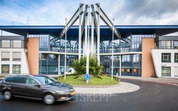 Kantoorpand gelegen op Eurodepark 1, Kerkrade, met moderne architectuur. Vooraanzicht toont grote glazen gevel met metalen constructie. Auto rijdt rond rotonde voor het gebouw.