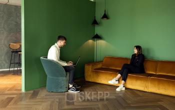 Moderne kantoorlounge met groene muren en houten vloer in Leeuwarden. Personen voeren zakelijke gesprekken op comfortabele stoelen en een bank. Ideale locatie voor kantoorruimte huren met een ontspannen sfeer.