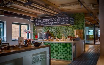 Kantine met kleurrijk betegelde muren en diverse eetstations in kantoorpand op Schipholweg 103, Leiden, waar mensen lunchen en werken.