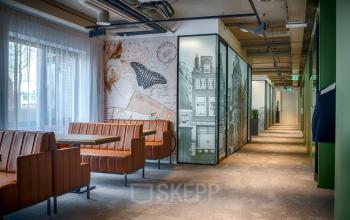 Kantoorruimte huren in modern gebouw aan de Schipholweg 103, Leiden. De ruimte heeft een lange gang met glazen wanden en comfortabele zitplaatsen aan tafel langs de muur.