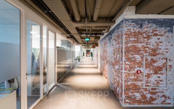 Moderne kantoorgang met glazen wanden in kantoorpand op Schipholweg 103, Leiden. Personen verrichten zakelijke activiteiten in diverse kantoren.
