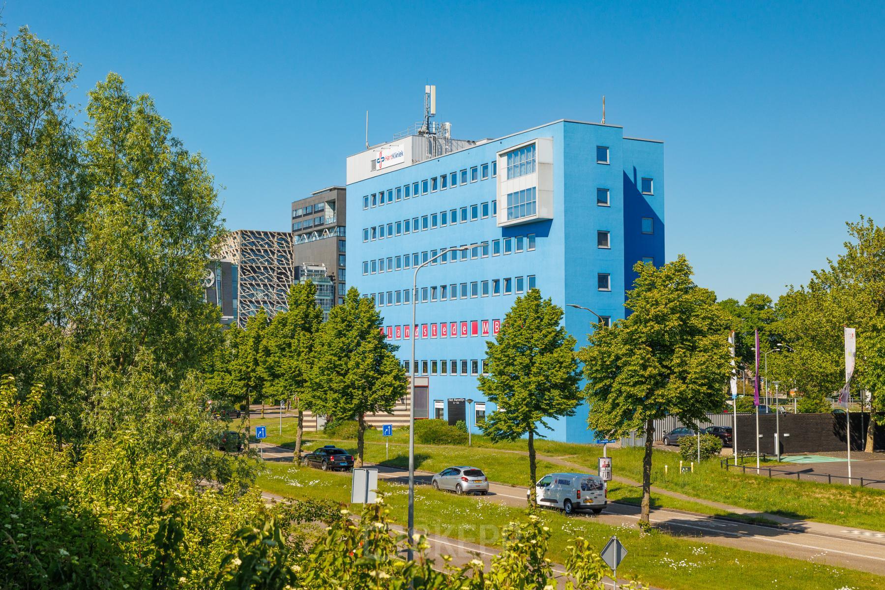 Vooraanzicht van het blauwe kantoorpand op Het Ravelijn 1-11, Lelystad, omgeven door bomen en een heldere lucht.