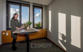 Binnen een kantoorpand in Leusden werkt een vrouw staand achter een bureau, met veel natuurlijk licht van de ramen. Kantoorruimte huren biedt hier een lichte en moderne ambiance.