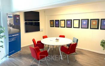 Vergaderruimte in kantoorpand op Wilhelminasingel 44-46 in Maastricht, met rode stoelen rond een witte tafel en kleurrijke kunst aan de muur. Ideaal voor kantoorruimte huren.