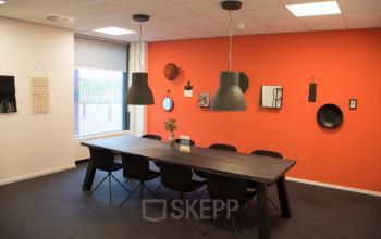Gedeelde vergaderruimte in het kantoorpand op Rijksweg 79 in Naarden, met een donkere houten tafel, acht zwarte stoelen, en een opvallende oranje muur met decoratieve elementen.