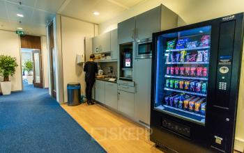 Moderne kantoorpand pantry op Jonkerbosplein 52 in Nijmegen met een medewerker die de koffiezetapparaat gebruikt en een drankenautomaat aan de rechterzijde. Comfortabel kantoorruimte huren in een levendige omgeving.