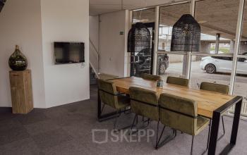 Kantoorruimte huren in Oosterhout Nb, Abdis van Thornstraat 74. Modern kantoorpandinterieur met houten tafel en groene stoelen, tegen een glazen wand met uitzicht op buiten. Neutrale tinten en stijlvolle verlichting.
