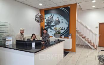 Entree van kantoorpand op Leeghwaterstraat 25, Reeuwijk met moderne receptiebalie waar twee mensen bezig zijn met ontvangst en zakelijke gesprekken. Lichte ruimte met subtiele decoratie.