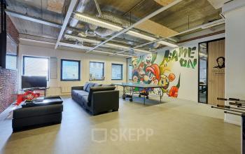Kantoorruimte met een speelse muur met graffiti en een tafeltennistafel, ideaal voor creatieve bedrijven om te huren. Het interieur is gemeubileerd met een bank en een voetbaltafel.