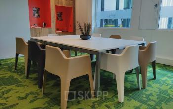 Een moderne kantooruimte met een lange tafel omringd door stoelen in een kleurrijke setting op Boven de Wolfskuil 3, Roermond, geschikt voor vergaderingen of als werkplek te huur.