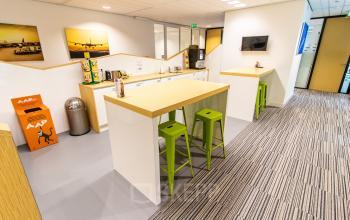 Lichte kantoorruimte in Rotterdam Airport met hoge tafels en groene krukken, ideaal voor pauzes. Er zijn ook kunstwerken aan de muur en een koffiemachine. Geschikt voor kantoorruimte huren.