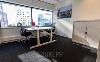 Stijlvol ingerichte kantoorruimte huren op Weena-Zuid 130, Rotterdam Centraal Station, met uitzicht op de stad door grote ramen. Twee bureaus met stoelen en een schilderij aan de muur.