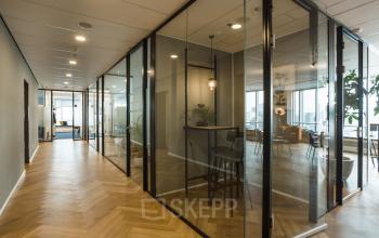 Moderne kantoorruimte te huur op Weena 690, Rotterdam Centrum. Glazen wanden scheiden de werkruimtes, met houten vloeren en verlichting. In de verte zicht op een open werkruimte met tafels en stoelen.