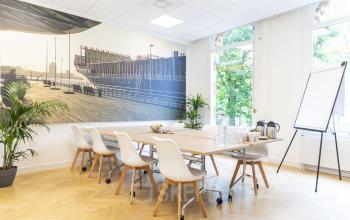 Kantoorruimte huren aan Westplein 12, Rotterdam Centrum. Modern ingerichte vergaderruimte met grote houten tafel, witte stoelen, flipover en planten. Grote ramen bieden natuurlijk licht.