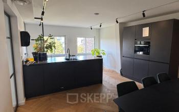 Moderne keuken in kantoorpand op Westplein 12, Rotterdam Centrum, met houten vloer, zwarte kasten en een ruime eettafel. Lichte ruimte met veel planten, geschikt voor vergaderingen of informele ontmoetingen.