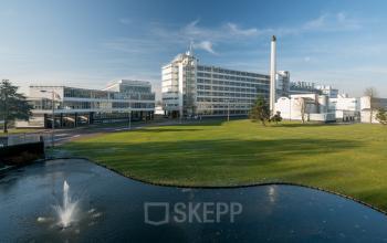 Kantoorpand op Van Nelleweg 1 in Rotterdam Delfshaven, omgeven door een groot groen veld met een fontein op de voorgrond. Ideale locatie voor het huren van kantoorruimte.