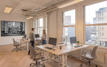 Lichte en ruime kantoor met houten vloer, grote ramen en bureaus met computers waar twee personen met elkaar werken of overleggen.