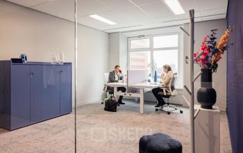 Twee mensen in gesprek in een lichte kantoorruimte op Wilhelminaplein 1-40, met modern meubilair en uitzicht op Rotterdam Kop van Zuid. Ideaal voor zakelijke ontmoetingen of vergaderingen.