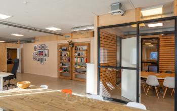 Moderne kantoorruimte huren aan de Linker Rottekade 292 in Rotterdam Noord met een open vloerplan, houten wanden en vernieuwende, lichte inrichting. Vergaderruimte met glazen scheidingswand zichtbaar in achtergrond.