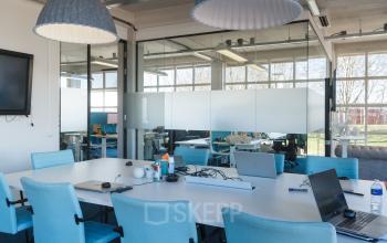 Vergaderruimte in kantoorpand op Overschieseweg 34 Schiedam met glazen wanden en blauwe stoelen rondom een tafel, laptops en kantoorbenodigdheden aanwezig.